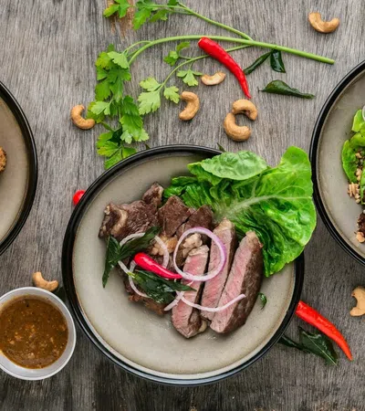 Three plates of gourmet dishes with grilled meat, fried appetizers, fresh herbs, lettuce, red chili, and cashew nuts on wooden table.