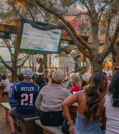 Outdoor sports bar with crowd watching a football game on multiple large screens under trees and string lights.
