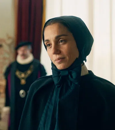 Woman in black medieval attire looking thoughtfully at an older man in a royal setting with candle and attendants.