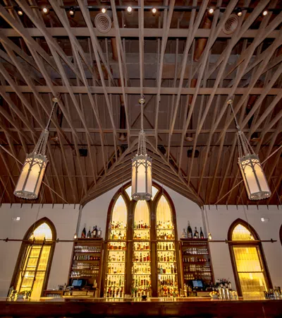 Upscale bar with gothic arched windows, illuminated shelves of liquor, and wooden ceiling beams with hanging lanterns.