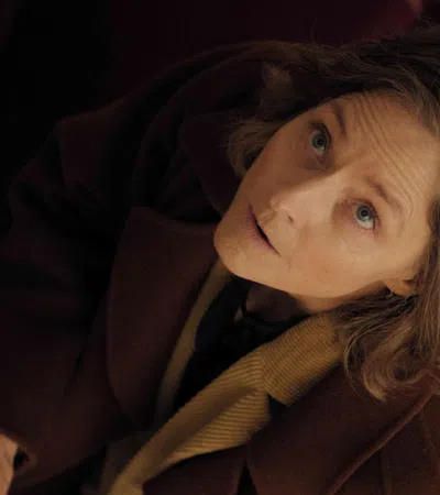 Woman in brown coat looking upward with a concerned expression in a dimly lit setting.