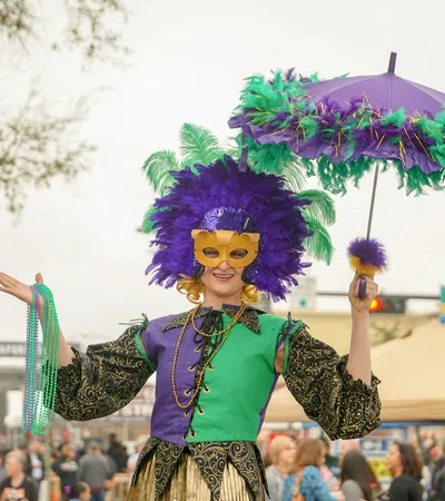Person wearing a colorful Mardi Gras costume with purple and green feathers, mask, beads, and decorated umbrella.