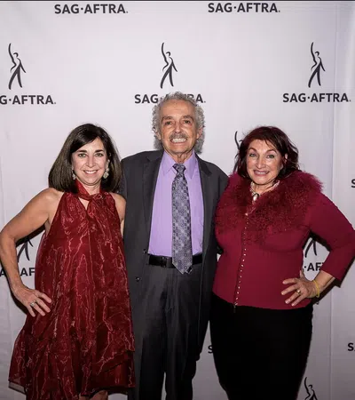 Three adults posing at SAG-AFTRA event with branded backdrop, two women in red outfits and one man in suit.