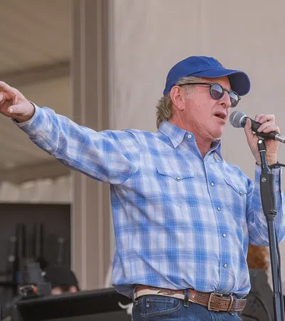 A man singing passionately on stage, wearing sunglasses and a blue shirt, with an audience in the background.