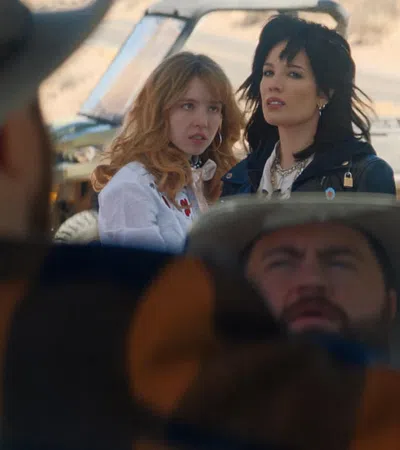 Two women stand outside a vehicle in a desert setting while a man in a hat is seated inside the vehicle.