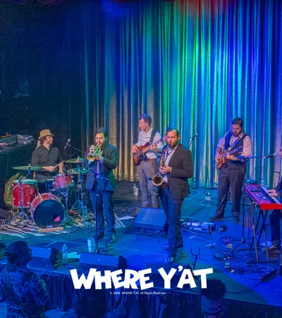 Jazz band performing live on stage with trumpet, saxophone, drums, guitar, keyboard, and bass under colorful lights.