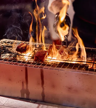 Flames flare up as meat pieces sizzle on a metal charcoal grill with a person grilling using tongs.