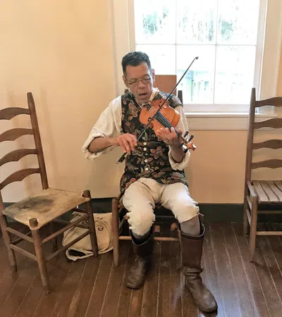 Man in historical costume playing violin while seated between wooden chairs on hardwood floor