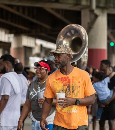 Man in orange shirt holding a cup with people and a sousaphone band under an overpass at a street event