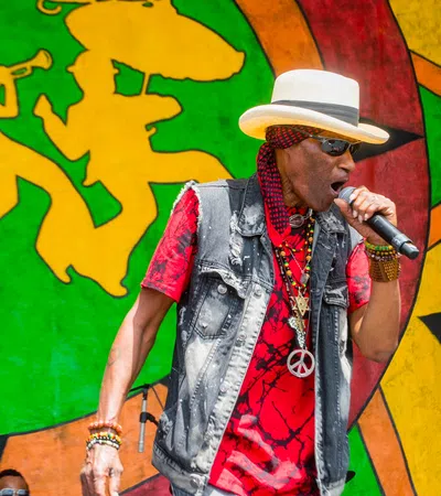 Male reggae singer wearing hat and sunglasses performs passionately on stage with colorful backdrop and band.
