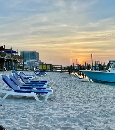 Empty blue and white beach chairs on sandy shore near docked boat at sunset with calm water and buildings.