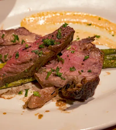 Grilled medium-rare steak slices served with asparagus and a creamy sauce on a white plate with herbs.