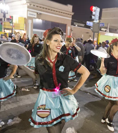 Women in vintage diner waitress costumes smiling and dancing with trays during a lively street parade at night.