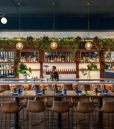 Stylish bar interior with wooden furniture, green plants, warm lighting, and a bartender preparing drinks behind the counter.