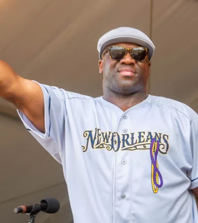Man in New Orleans jersey and sunglasses holds microphone on stage under tent ceiling