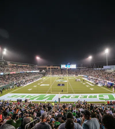 Nighttime football game with full stadium and bright floodlights illuminating the field and crowd.