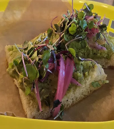 Open-faced sandwich with guacamole, pickled red onions, and microgreens on focaccia bread in a yellow basket.