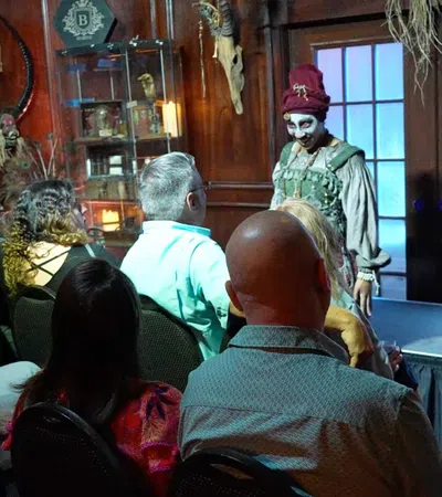 Audience watches performer in creepy costume and makeup on stage in dimly lit room with spooky decorations.