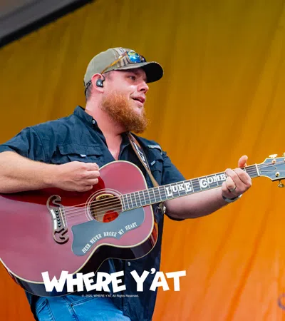 Male country musician playing guitar on stage with a yellow backdrop and wearing a cap and sunglasses