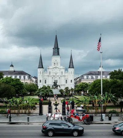 St. Louis Cathedral in New Orleans with horse carriages, cars, American flag, and cloudy sky in the background