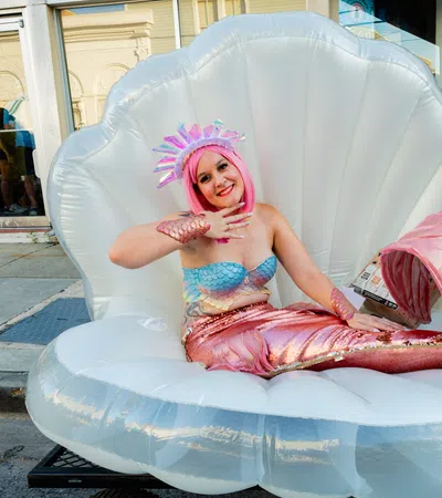 Smiling woman in a pink mermaid costume sitting on a large inflatable clam shell on a city street.