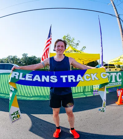 Runner celebrates victory holding Orleans Track Club banner at outdoor race finish line on sunny day