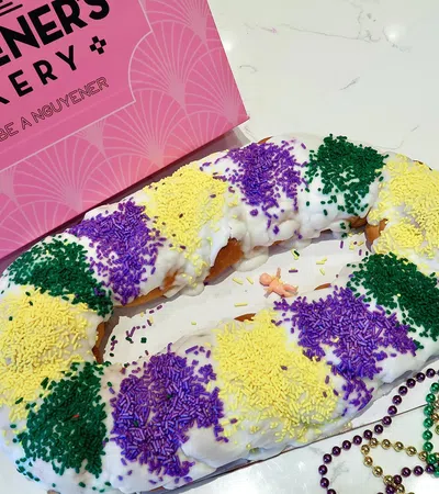 King cake with white icing and purple, yellow, green sprinkles next to pink Douceur Guyenere's Bakery box