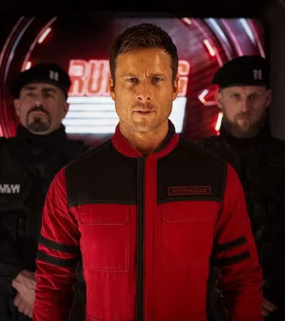 Man in red and black jacket with two guards in black uniforms standing behind him under dim lighting.