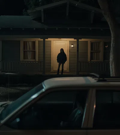 Person standing in the lit doorway of a house at night with a parked car in the foreground.