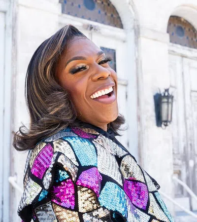 Smiling person in colorful sequin jacket posing outdoors near weathered white doors and stone steps