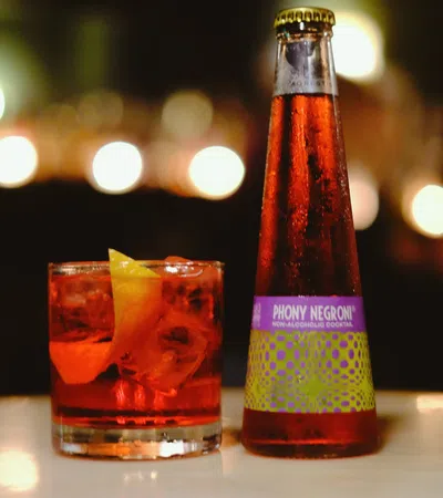 Bottle of Phony Negroni non-alcoholic cocktail next to a glass with ice and orange peel on a table.