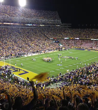 Nighttime college football game at LSU stadium with packed stands and fans waving towels.