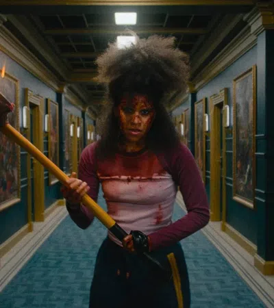 Person with blood-stained clothes holding a flaming torch standing in a dimly lit hallway lined with paintings.