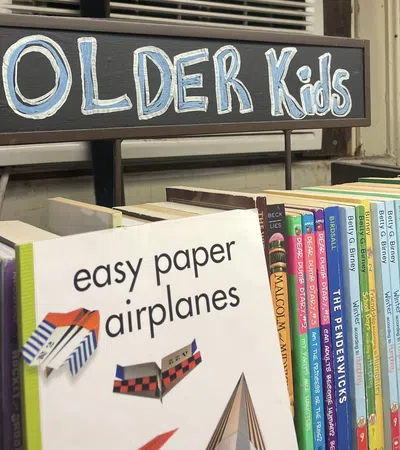 Books for older kids displayed on a shelf with a sign reading Older Kids and a book about easy paper airplanes.