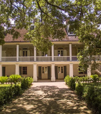 Historic two-story plantation house surrounded by lush greenery and shaded by large oak trees.