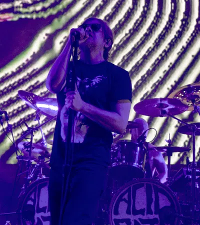 Male rock vocalist singing on stage with microphone and drummer performing behind during purple light concert.