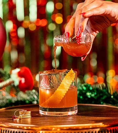 Hand pouring a festive cocktail with an orange twist on a wooden table surrounded by Christmas ornaments and lights.