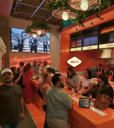 Busy Mexican restaurant with neon sign, orange tiled bar, hanging plants, and customers enjoying food and drinks.