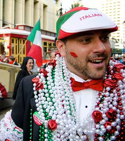 Two men wearing festive Italian-themed beads and accessories during a street celebration with flags and trolley.