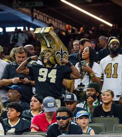 Fans wearing New Orleans Saints jerseys and gear cheer in a crowded stadium during a football game.