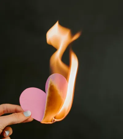 Hand holding a pink paper heart burning with bright orange flames against a dark background.