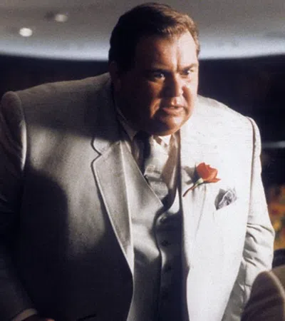 Man in a light suit with a rose boutonniere talks intensely to seated men in a dimly lit restaurant setting.
