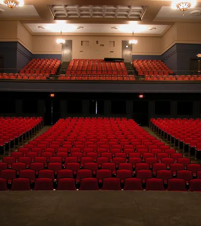 Empty theater with rows of red seats and a balcony, viewed from the stage, under warm lighting.