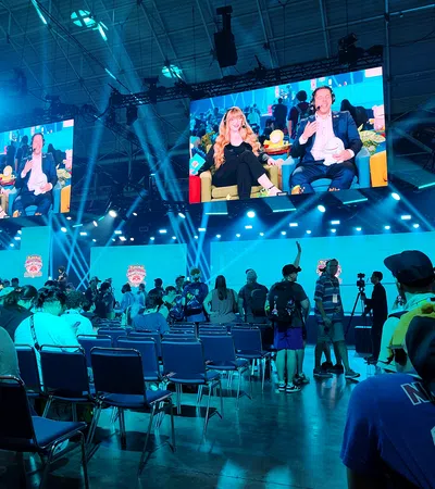 Crowded gaming event with audience watching live commentators on large screens under blue stage lighting.