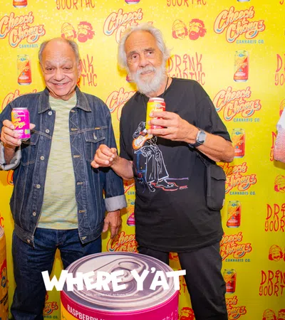 Two men smiling and holding cans of Cheech & Chong's cannabis drink in front of a branded yellow backdrop.