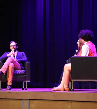 Man and two women seated on stage in armchairs having a discussion with microphones under purple stage lighting.