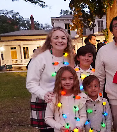 Family dressed in holiday lights standing outdoors near a giant on-off switch decoration at dusk