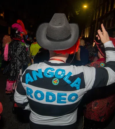 Person wearing Angola Rodeo striped shirt and cowboy hat at a lively nighttime costume event with colorful attendees.