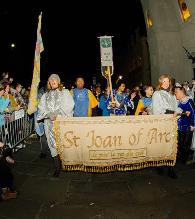 Night parade with people dressed as St Joan of Arc and medieval figures holding a banner and flags in front of a crowd.