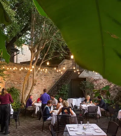 Outdoor restaurant patio with diners, string lights, brick walls, and tropical leaves creating a cozy ambiance.